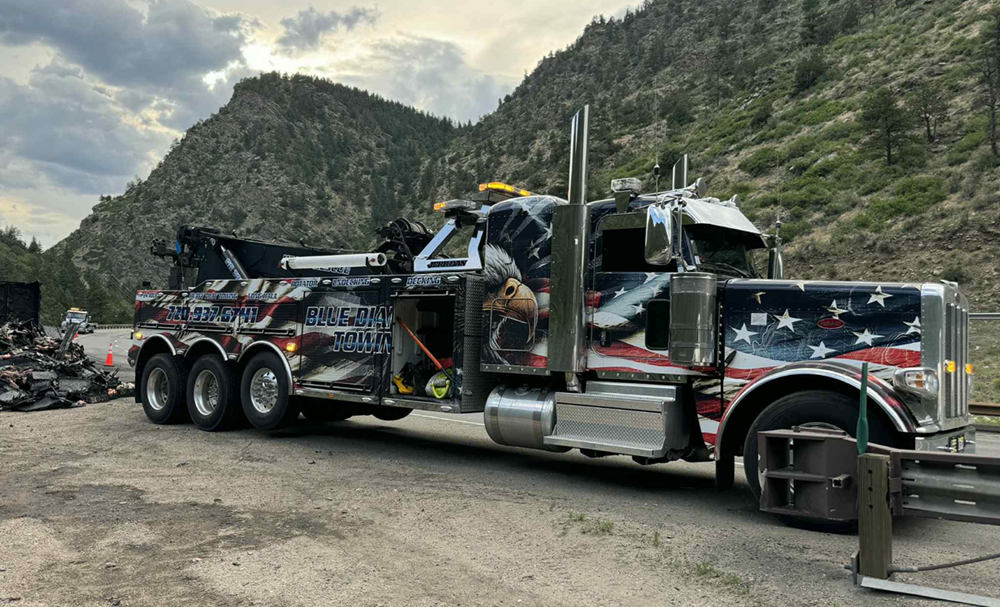 Patriotic tow truck with eagle graphic parked on roadside near mountains.