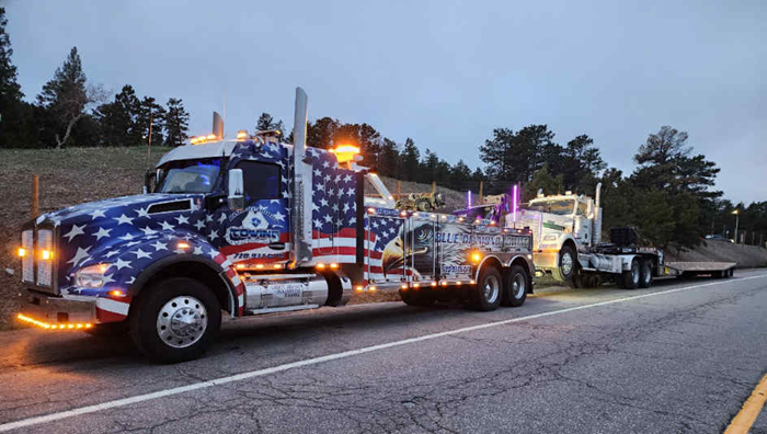American-flag-wrapped tow truck towing another truck on a road at dusk, lights on.