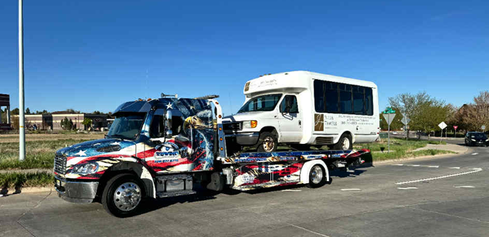 Tow truck with American flag design carrying a white passenger van on a sunny day.