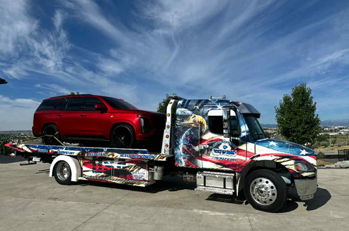 Tow truck with American flag-themed graphics carrying a red SUV on a sunny day.