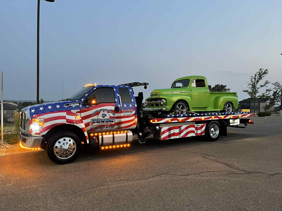 Tow truck with American flag design carrying a green vintage pickup truck.