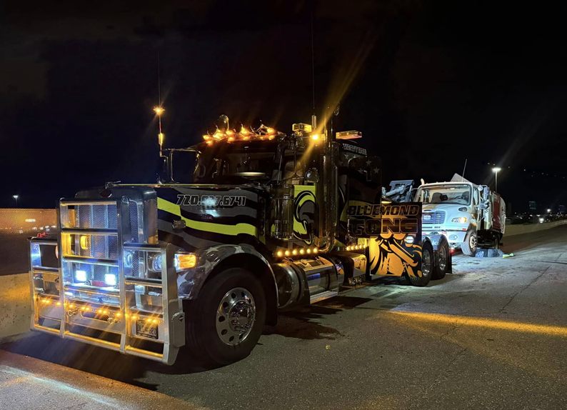 Semi-truck towing a damaged garbage truck at night; both are illuminated.