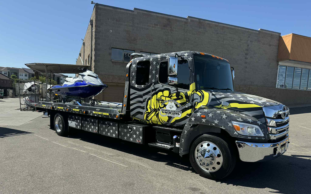 Tow truck with jet ski, in front of a brick building, with patriotic themed decals on the truck.