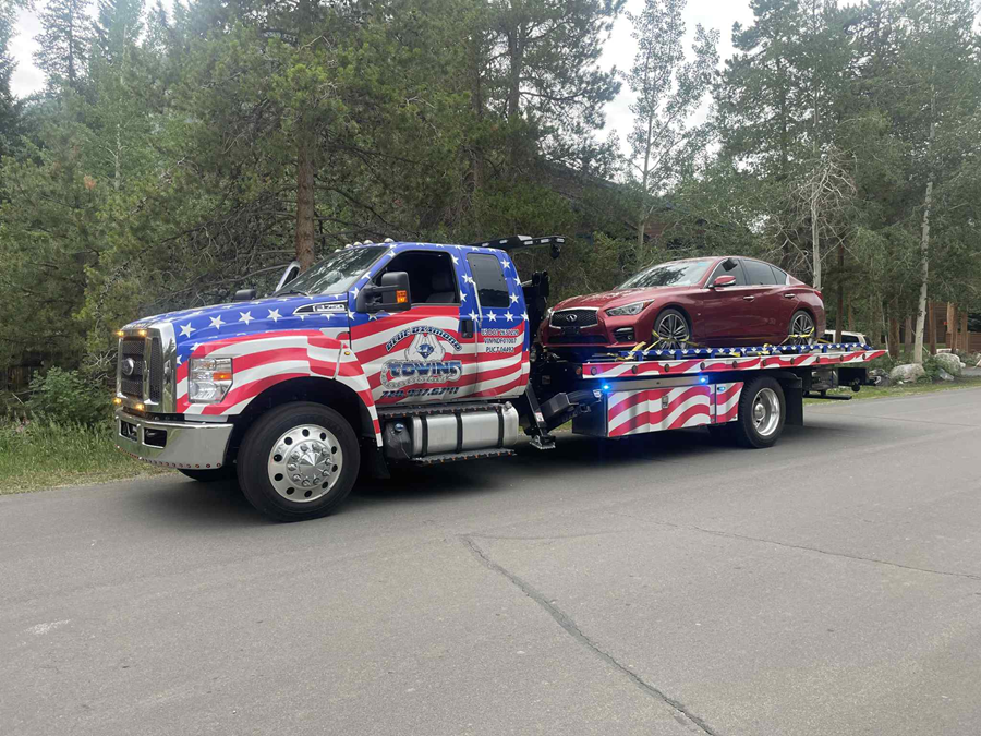 Tow truck with American flag design carrying a red car on a road, trees in the background.