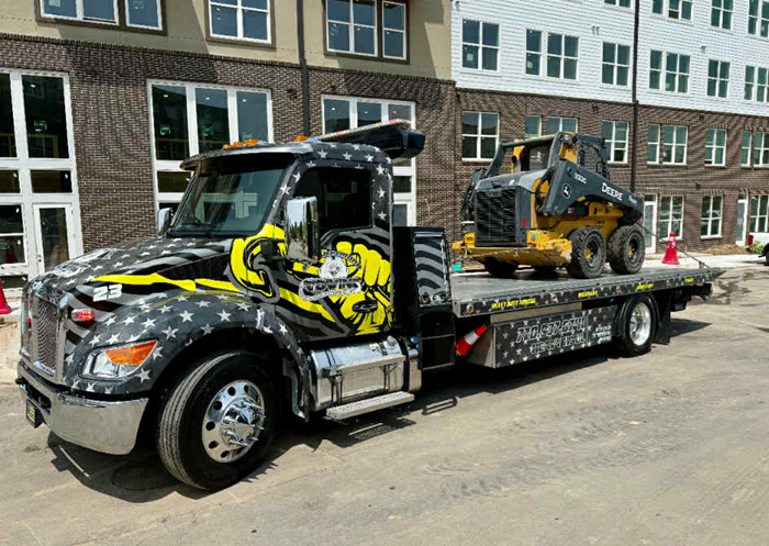 Flatbed tow truck with yellow construction equipment on its bed. Decorated with stars and stripes.