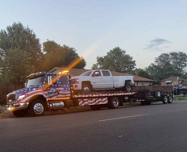 Tow truck with American flag design hauling a white pickup truck on a trailer.