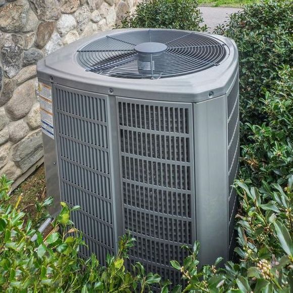 A large air conditioner is sitting in a bush next to a stone wall.