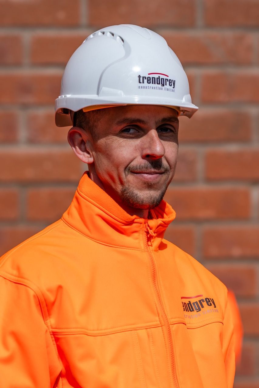 A man wearing a hard hat and an orange jacket is standing in front of a brick wall.