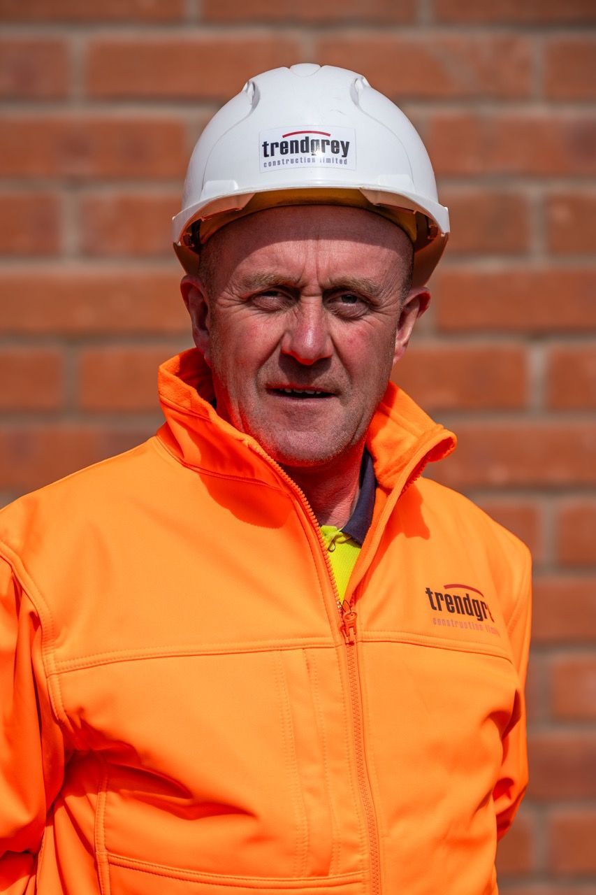 A man wearing a hard hat and an orange jacket is standing in front of a brick wall.