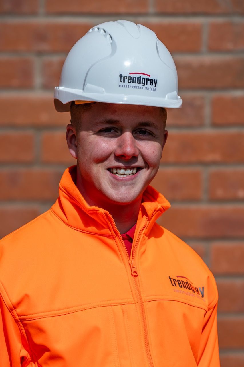 A man wearing an orange jacket and a white hard hat with trinity on it