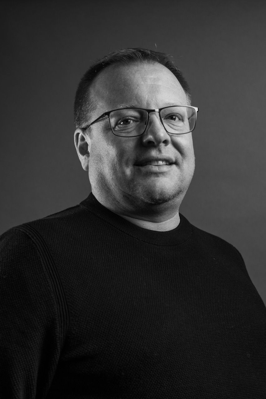 Nick Henderson, a man wearing glasses and a black sweater is smiling in a black and white photo.