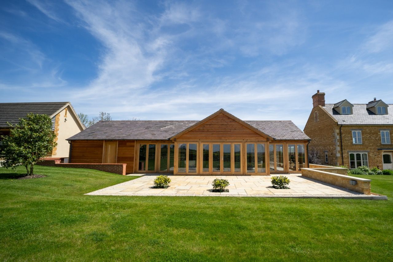 St Dennis Pool House, A large wooden house with a lot of windows is sitting in the middle of a lush green field.