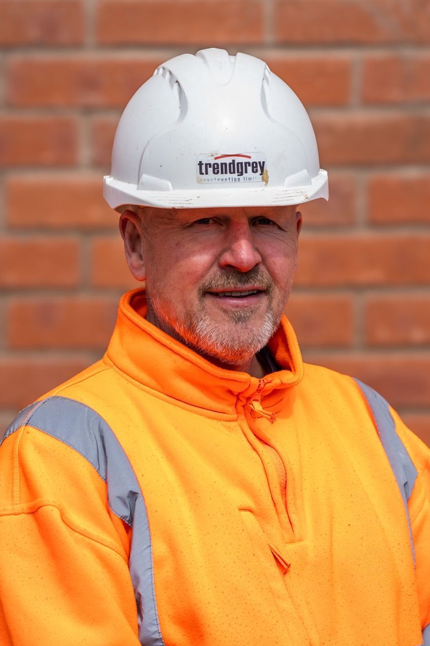 A man wearing a hard hat and an orange jacket is standing in front of a brick wall.