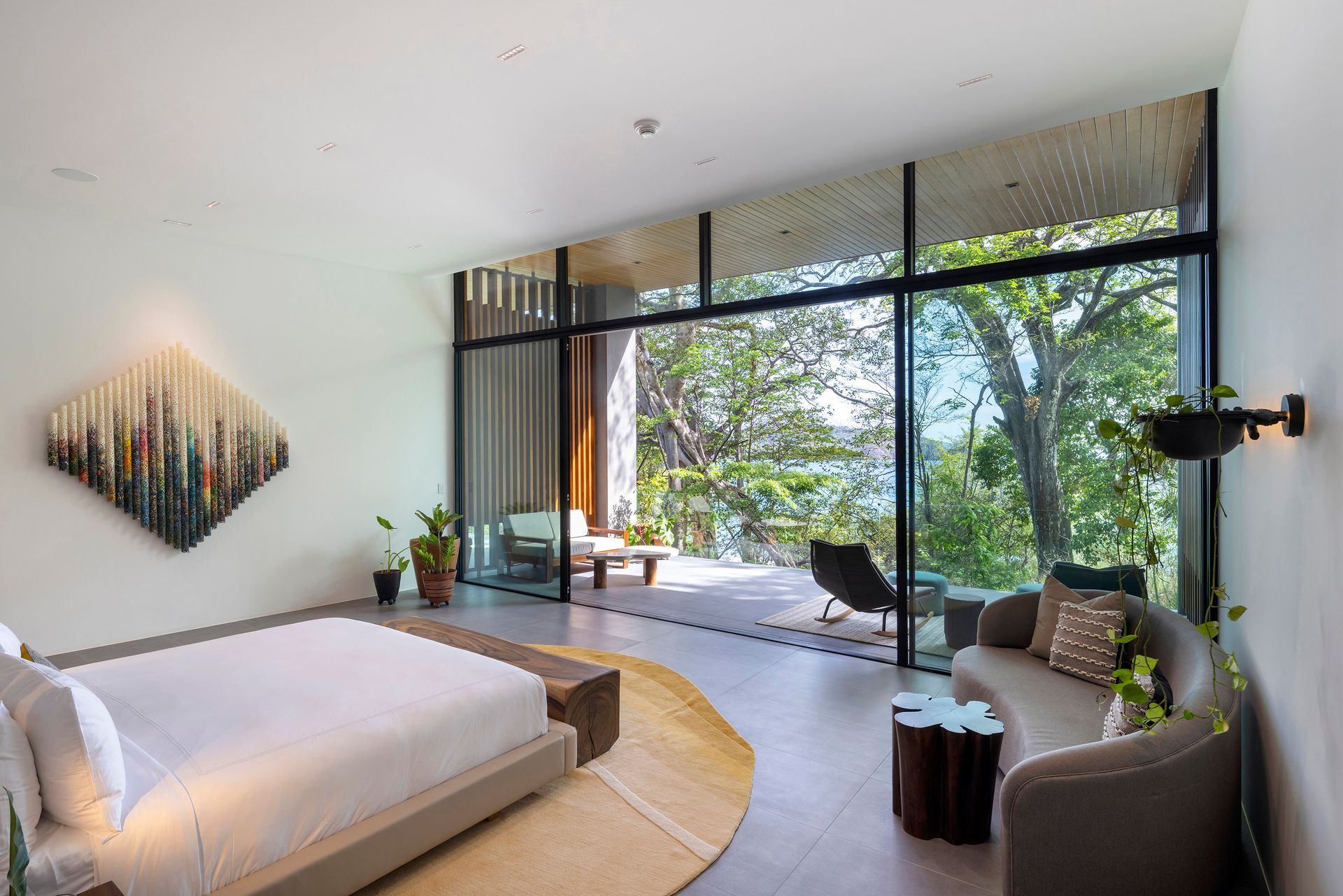Modern bedroom with large window overlooking greenery and a sitting area on a deck.