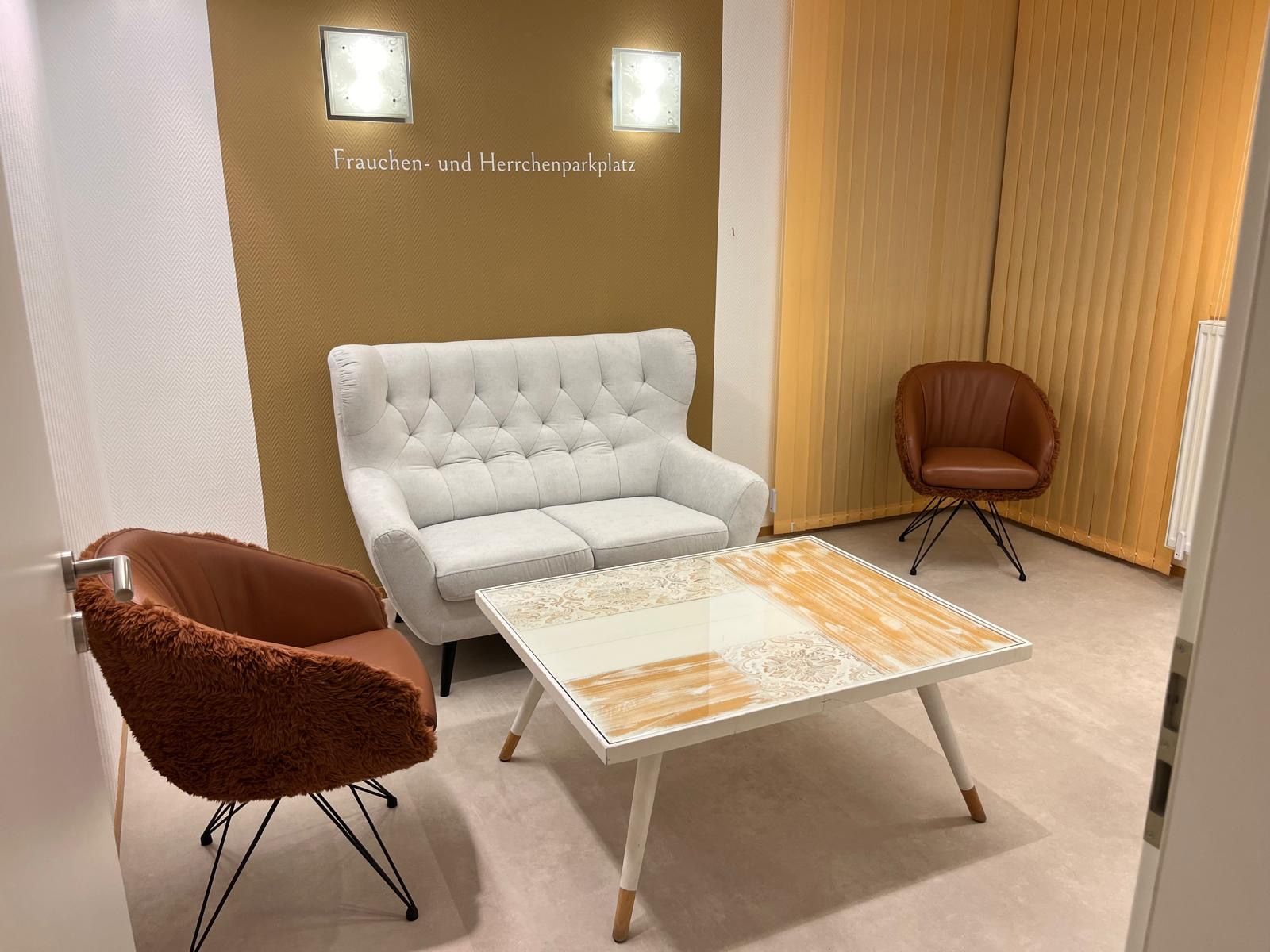 Gemütliches Wartezimmer mit weißem Sofa, zwei braunen Stühlen und einem Mosaik-Couchtisch; goldene Wand.