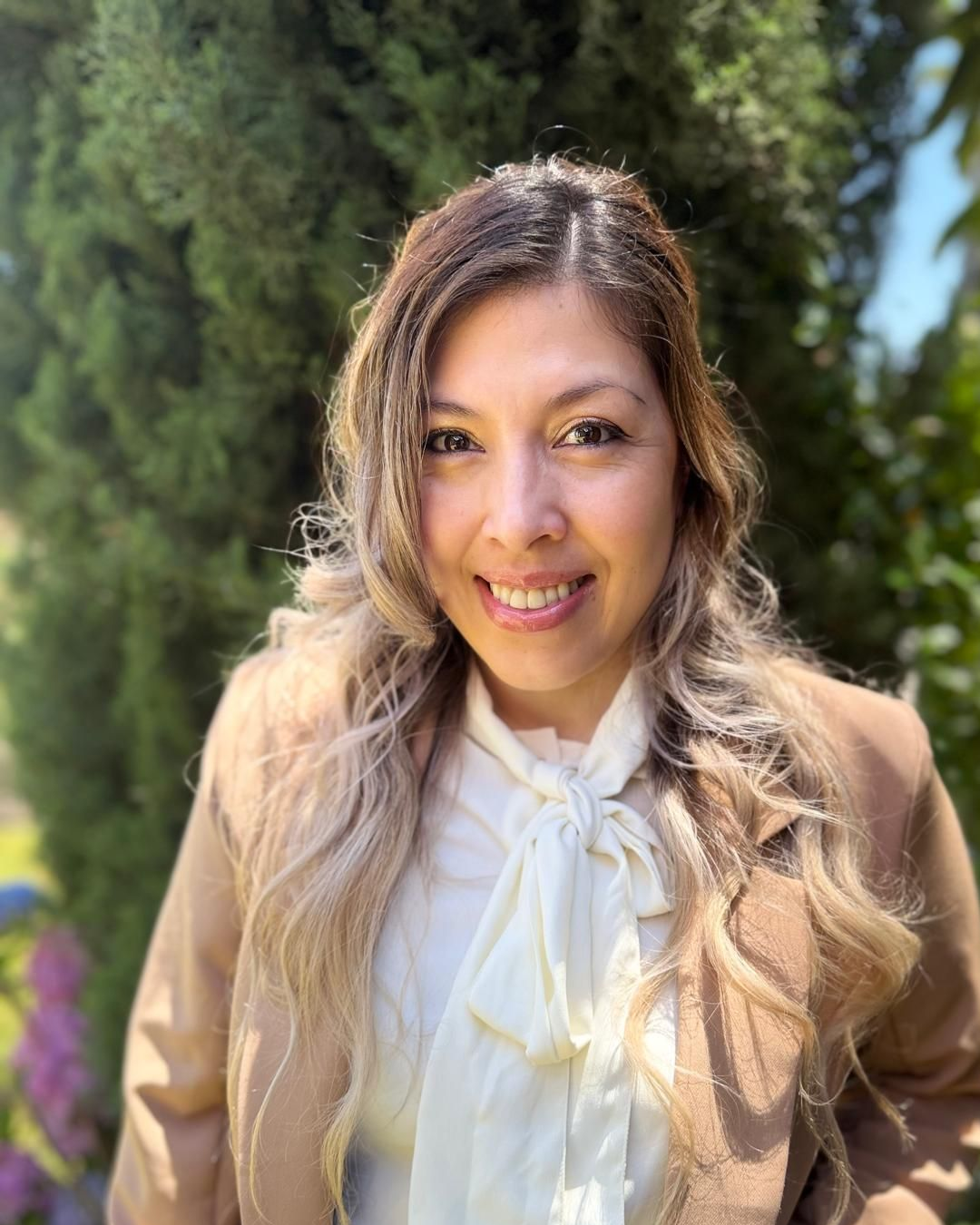 Woman with long highlighted hair smiles in a light blue striped button-up shirt outdoors.
