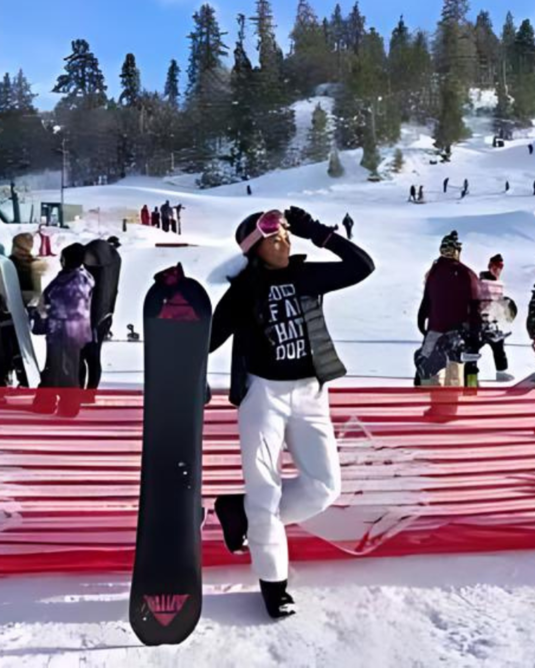 Person holding a snowboard poses in the snow, wearing white snow pants and a black top; ski resort in the background.