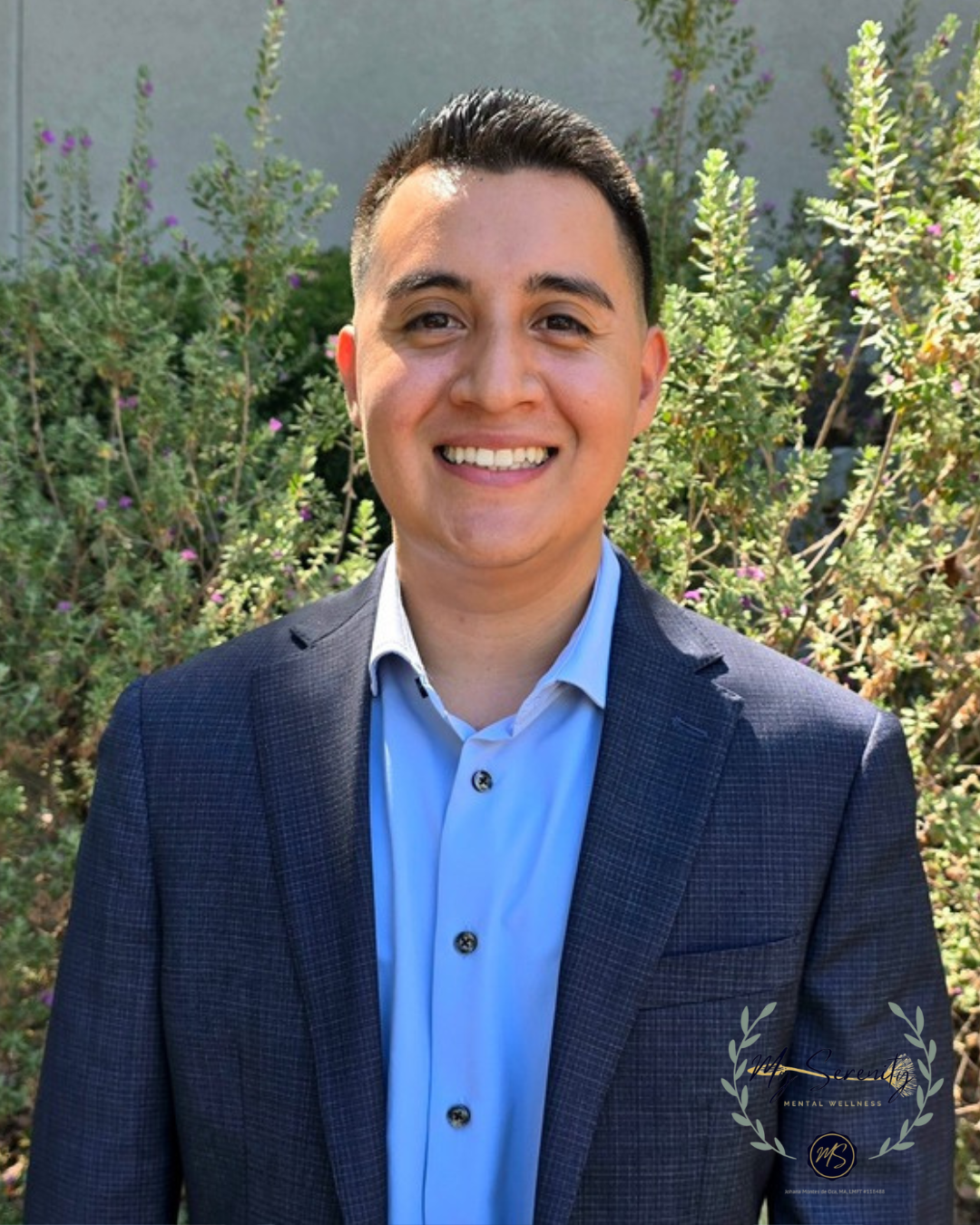 Smiling person in a navy blazer and light blue shirt standing outdoors in front of greenery.