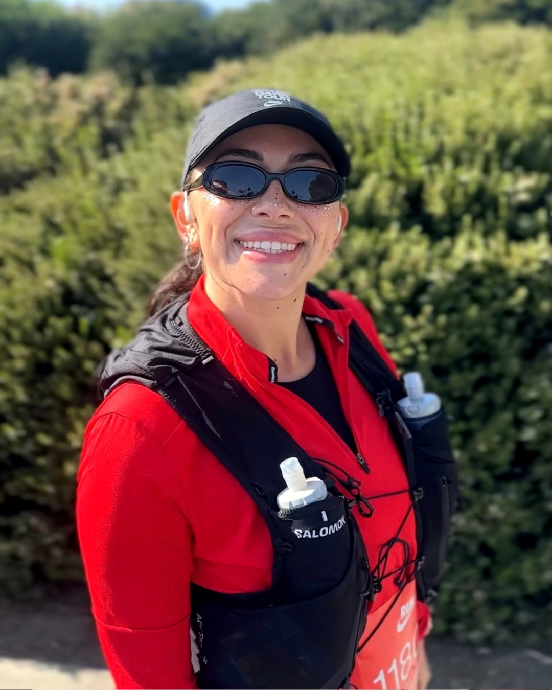 Person holding a snowboard poses in the snow, wearing white snow pants and a black top; ski resort in the background.