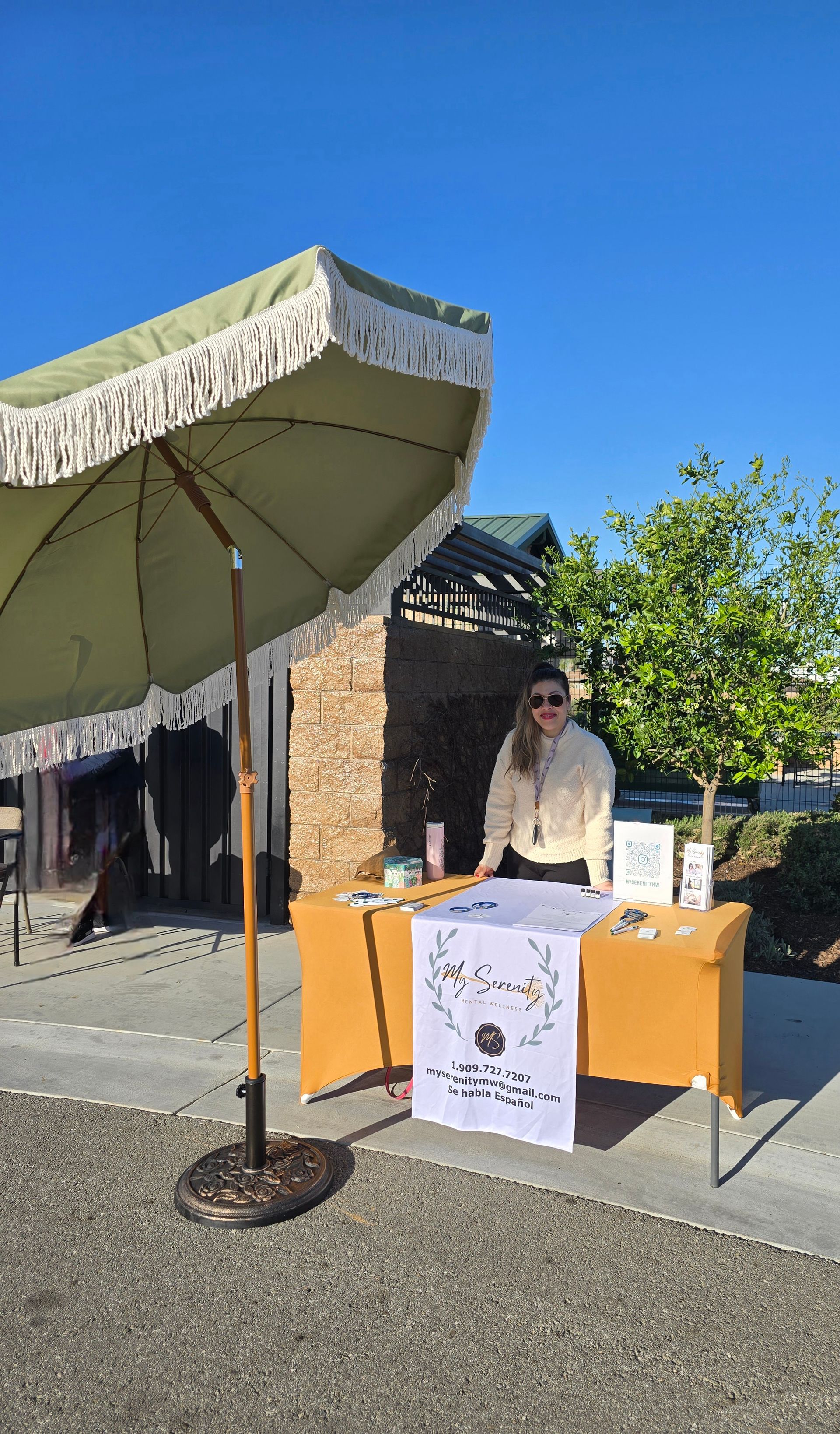 Woman stands behind a table under a fringed umbrella. Display of items on the table, clear blue sky.