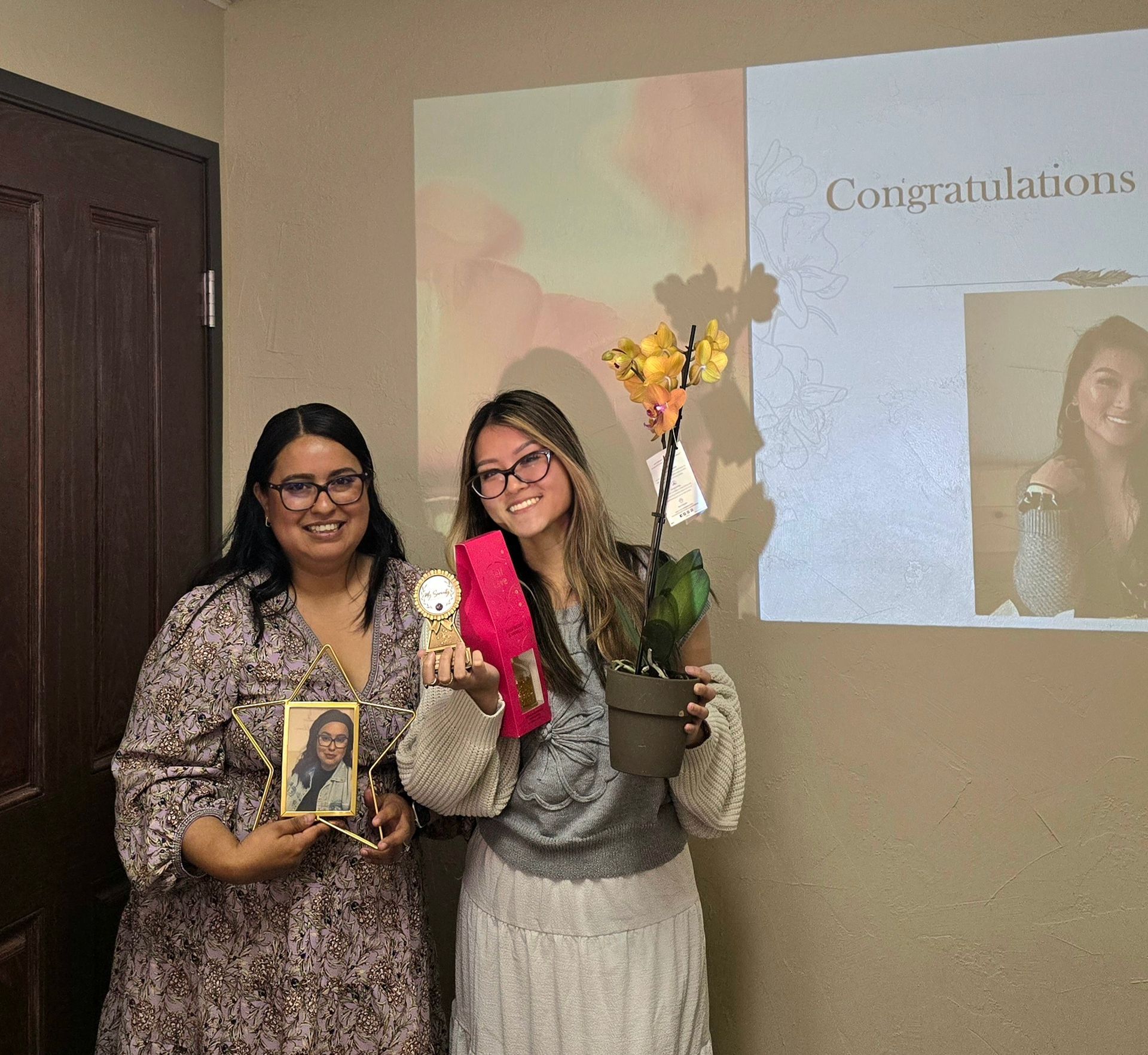 Two women smile, holding gifts: a framed photo, a flower, and a ribbon. 