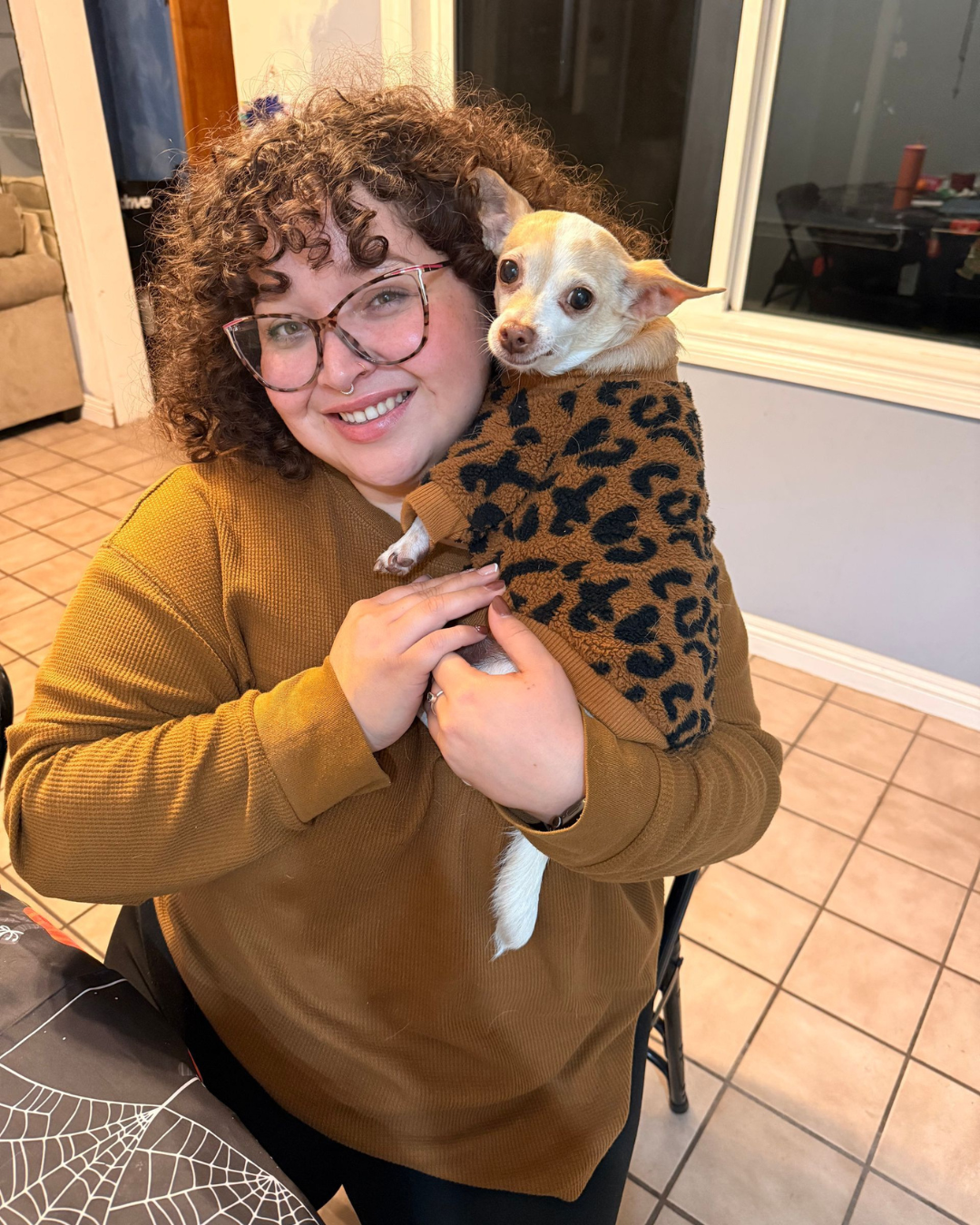 Person holding a snowboard poses in the snow, wearing white snow pants and a black top; ski resort in the background.