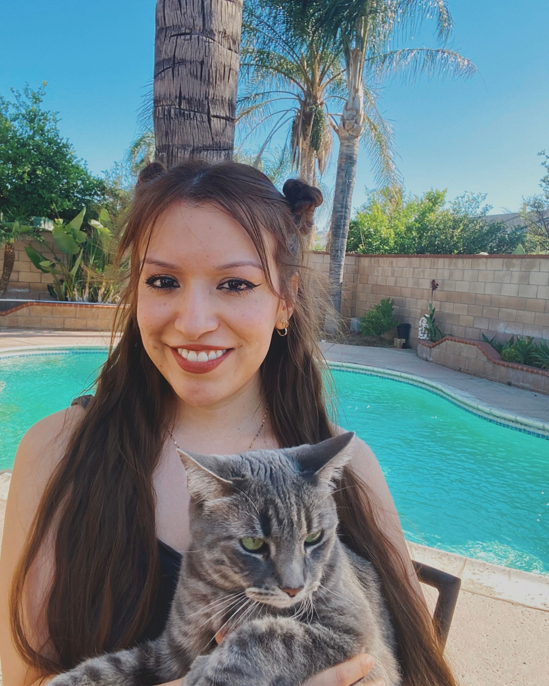 Person holding a snowboard poses in the snow, wearing white snow pants and a black top; ski resort in the background.