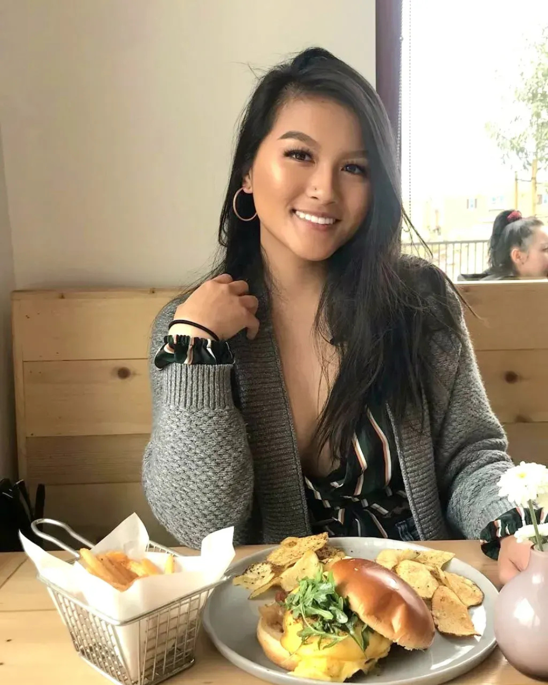 Woman smiling at a table, eating a burger and chips in a restaurant, wearing a sweater.