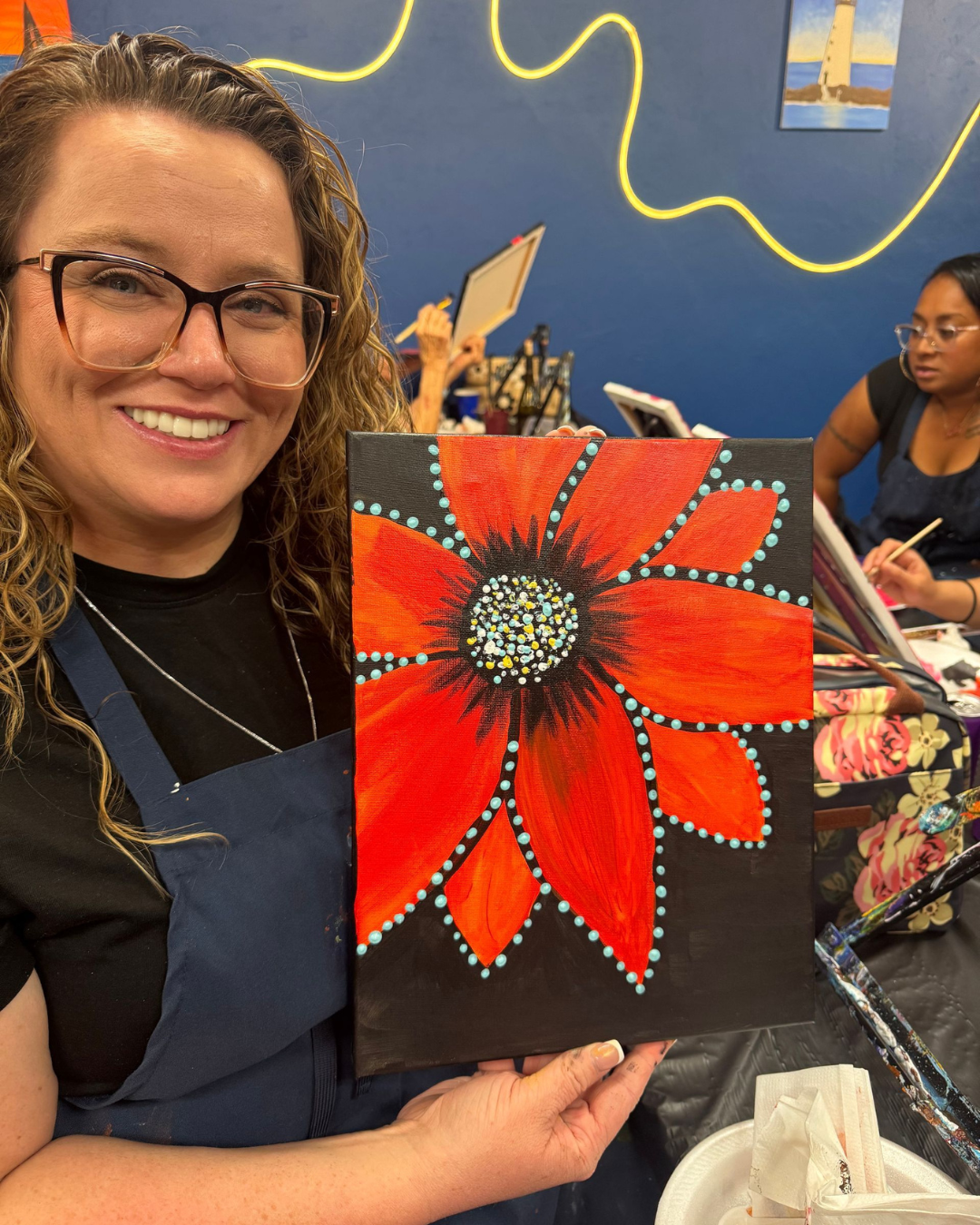 Person holding a snowboard poses in the snow, wearing white snow pants and a black top; ski resort in the background.
