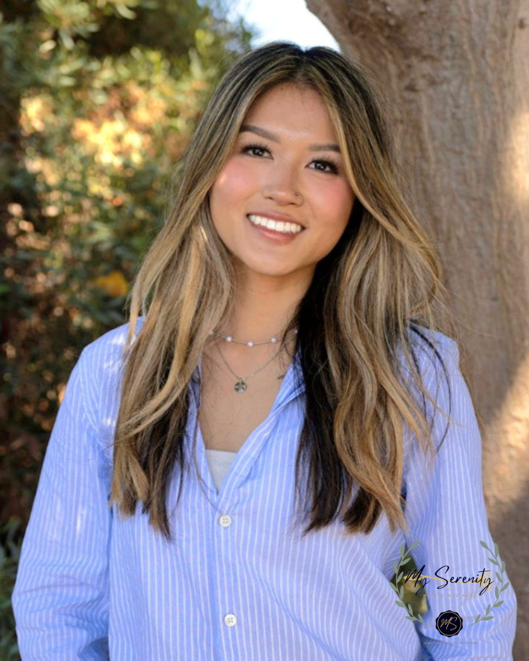 Woman with long highlighted hair smiles in a light blue striped button-up shirt outdoors.