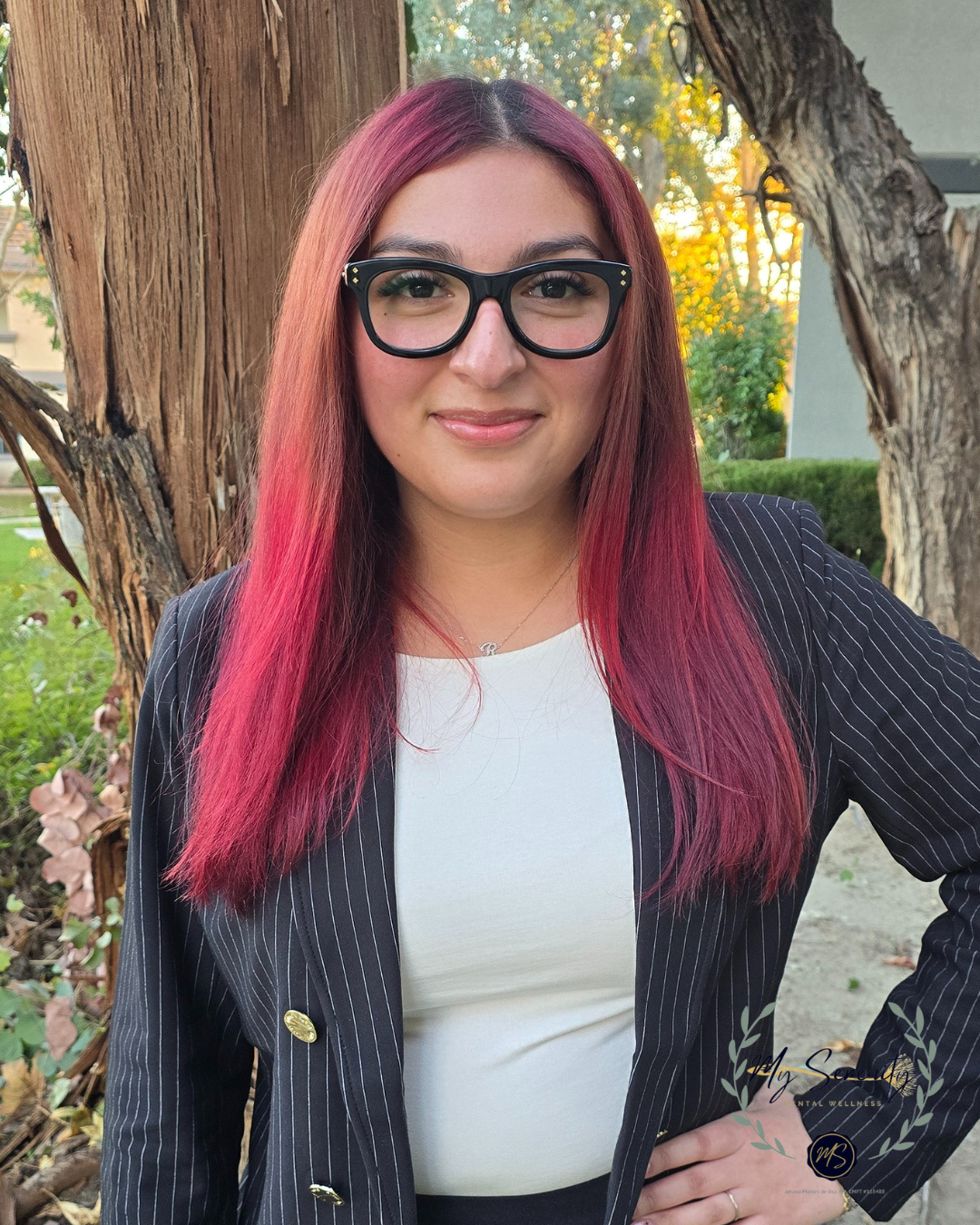Woman with pink-red hair, glasses, and a black blazer, smiles in front of a tree.