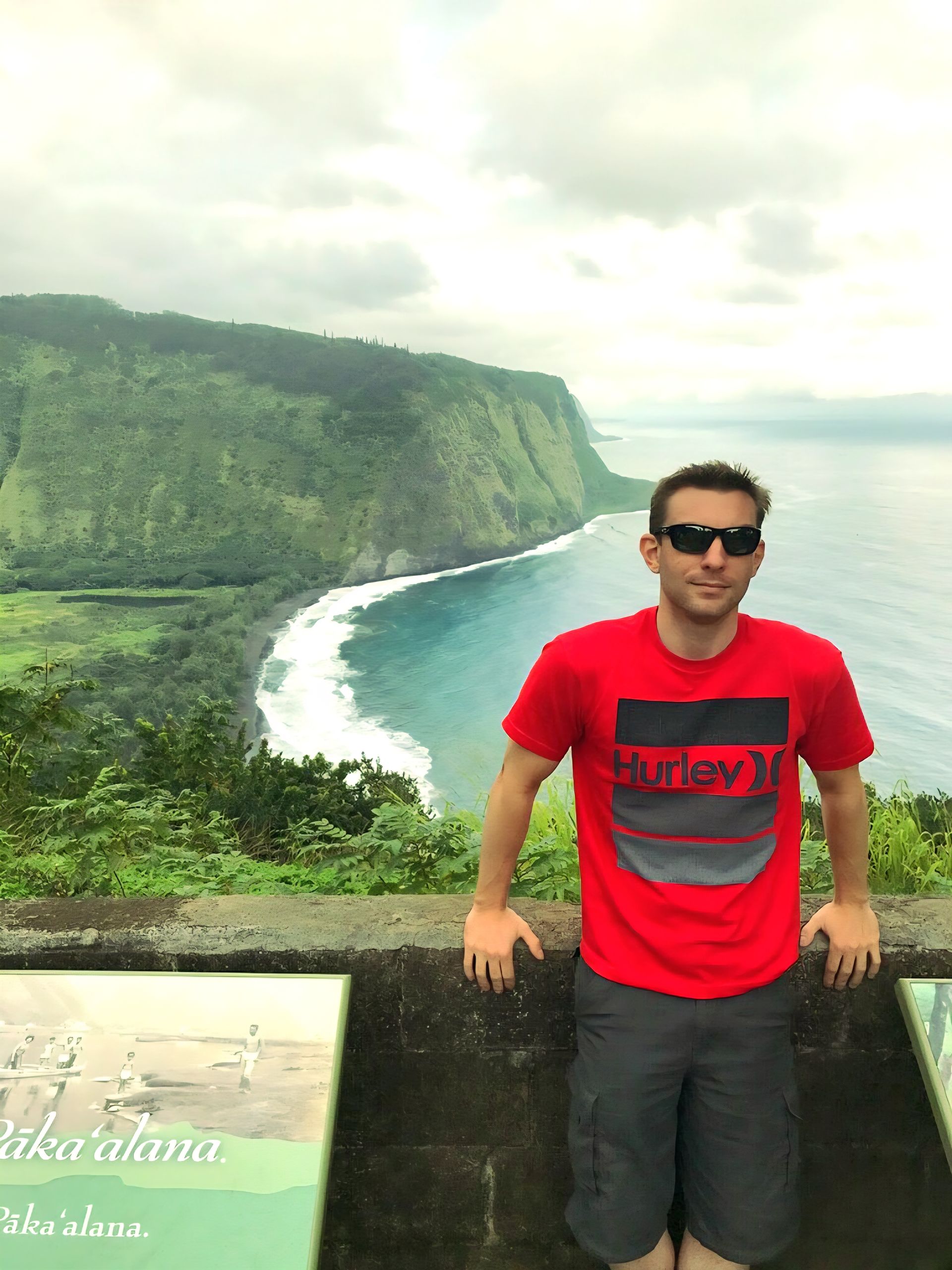 Man in red shirt and sunglasses poses at Waipio Valley overlook. Ocean and green cliffs visible.