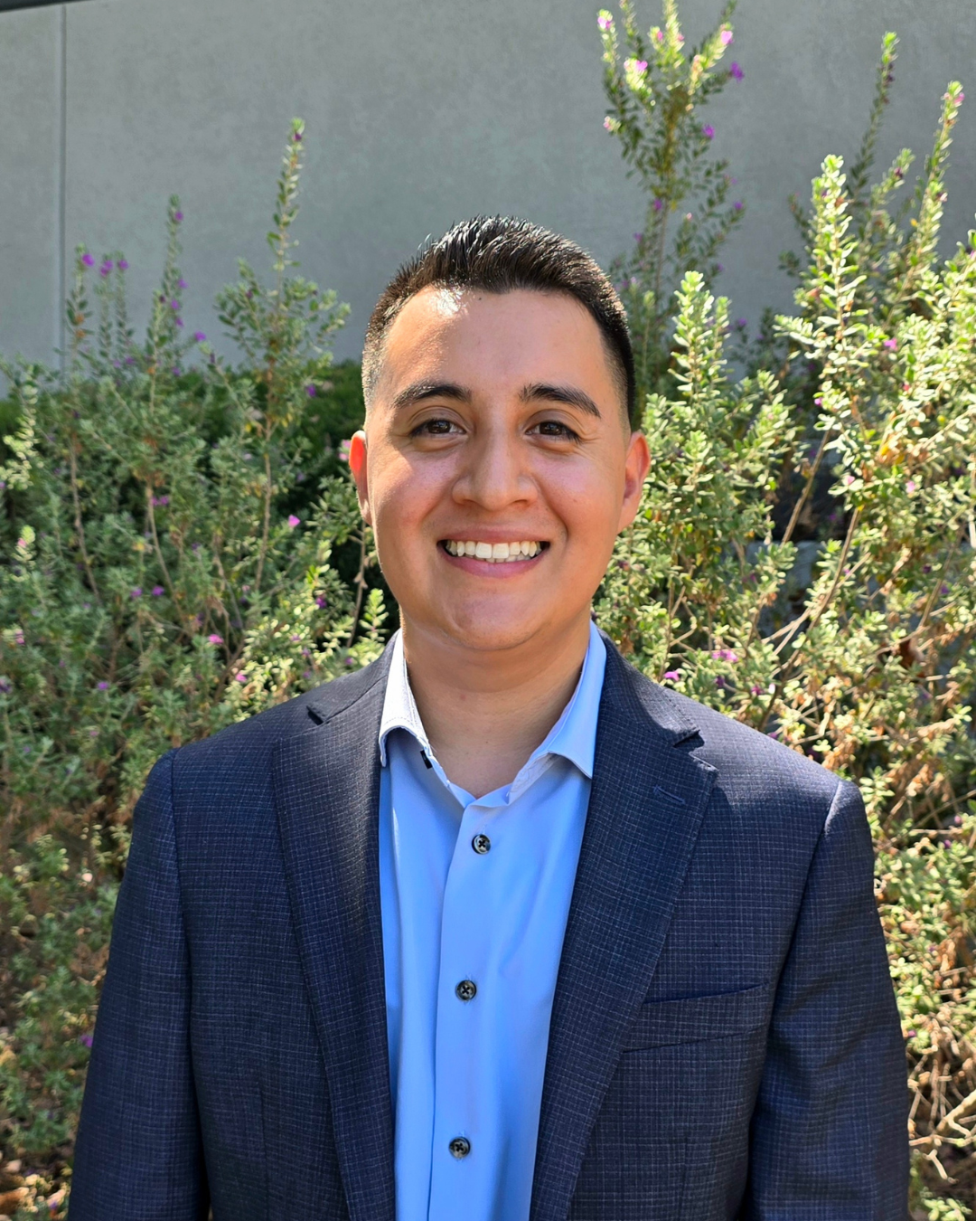 Smiling person in a navy blazer and light blue shirt standing outdoors in front of greenery.