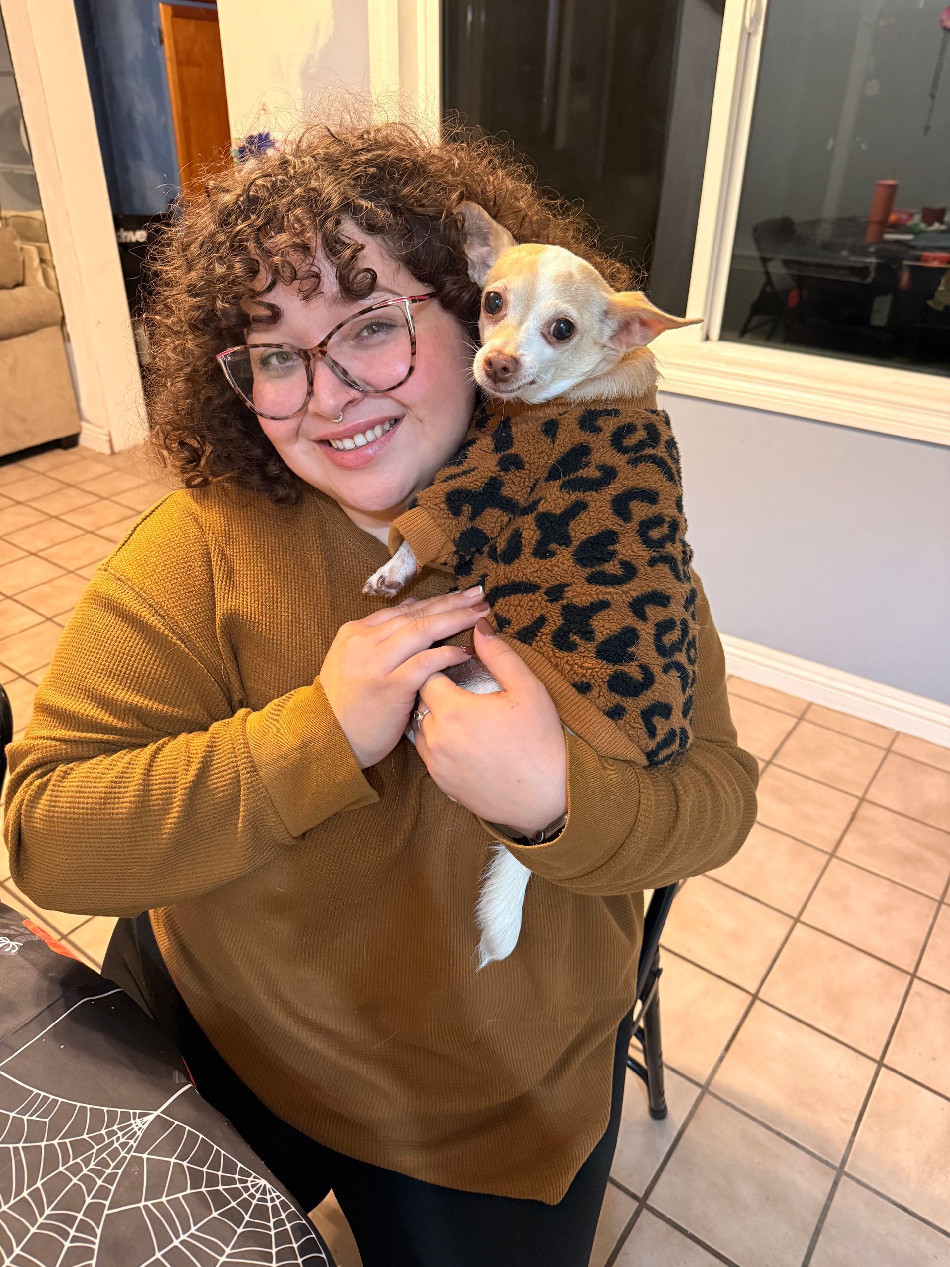 Person smiling, holding a small dog wearing a leopard print sweater. They're indoors.