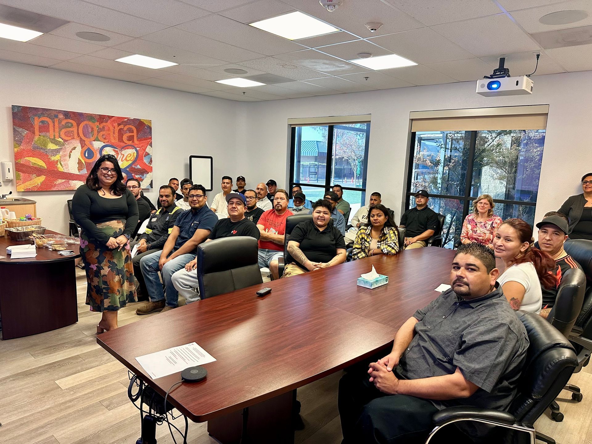A woman speaks in a conference room to a seated group. Artwork on the wall; window in the background.