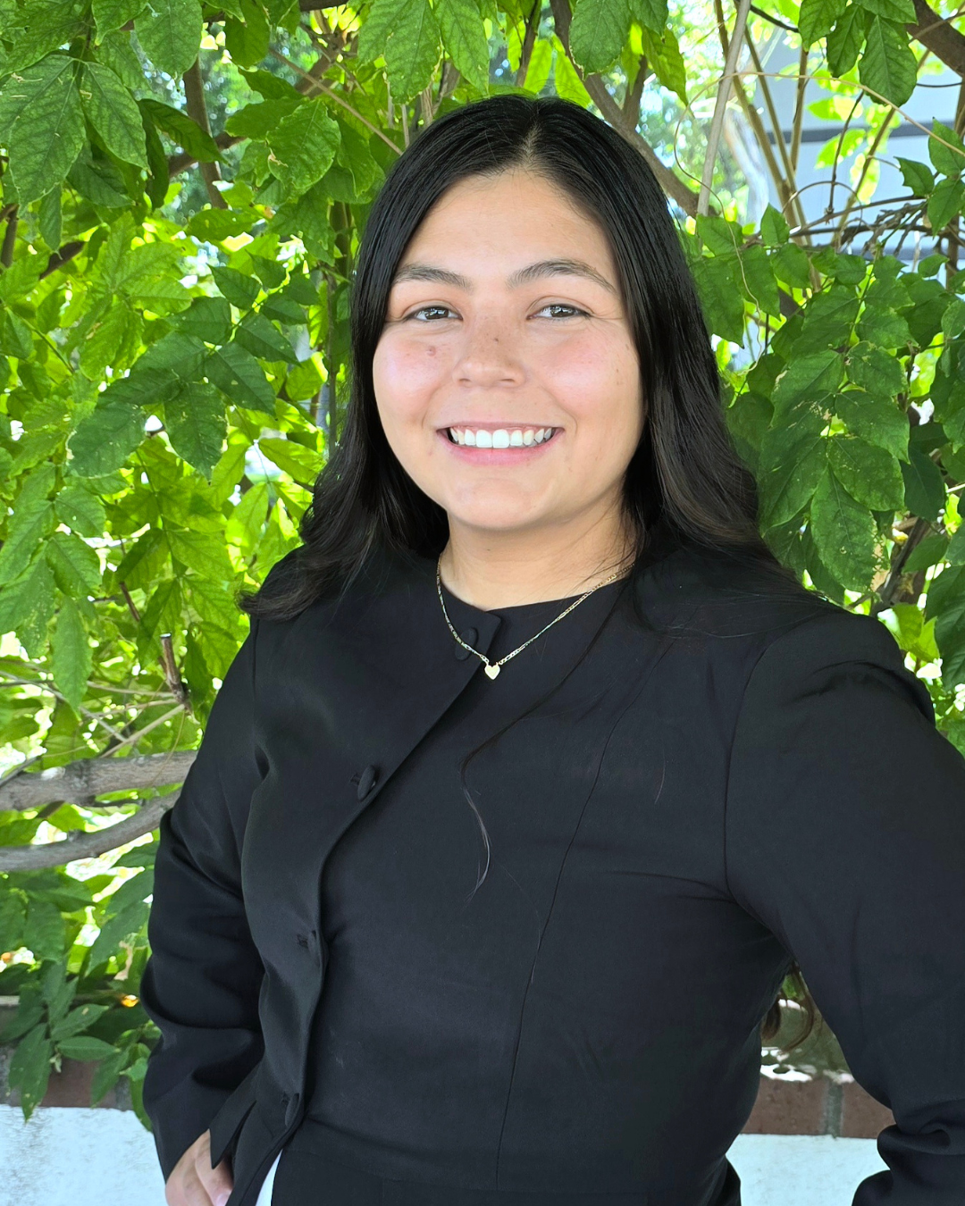 Smiling person in a black top standing outdoors in front of green leaves.