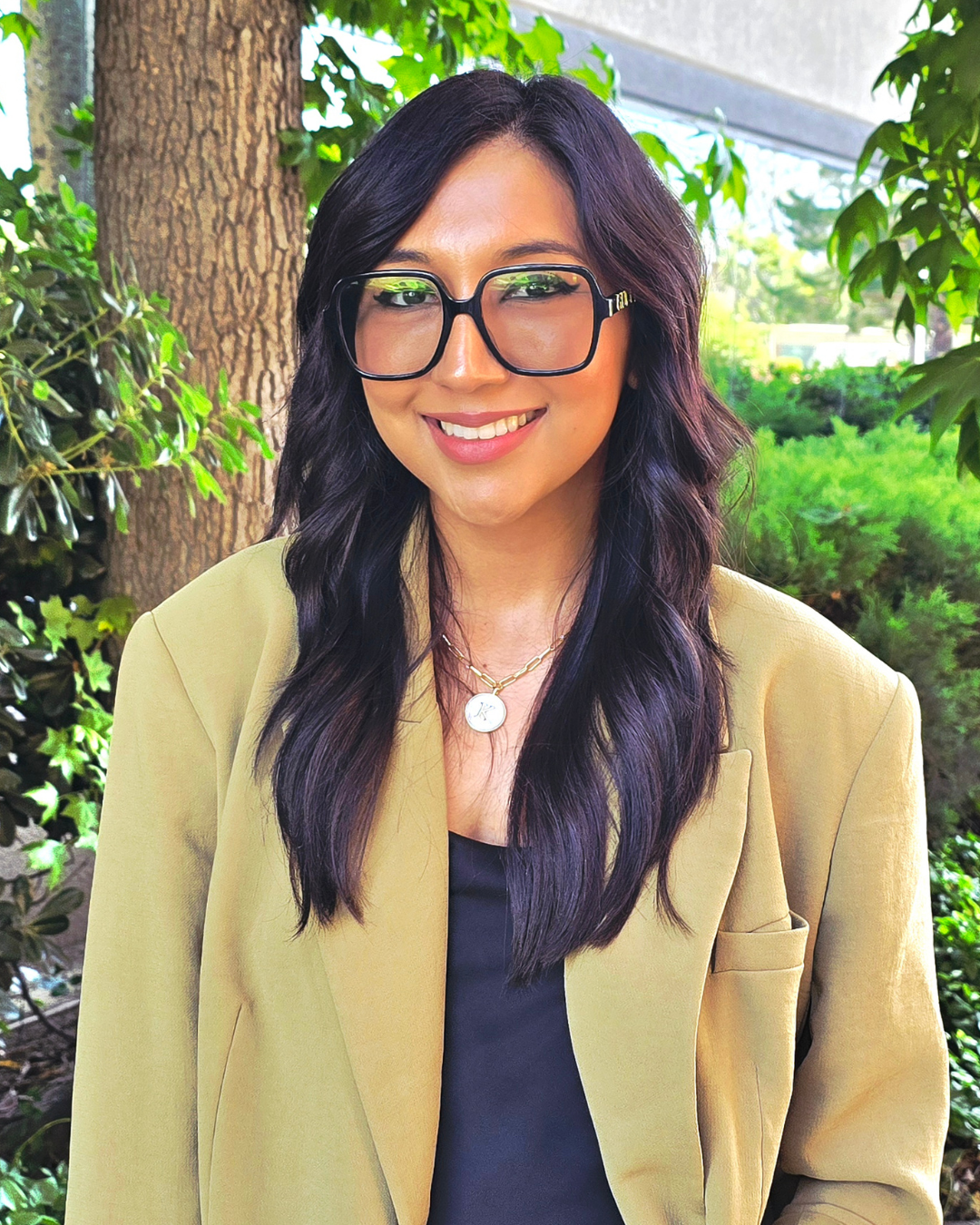 Woman with dark hair and glasses smiles, wearing a tan blazer and black top, standing outside a building near a tree.