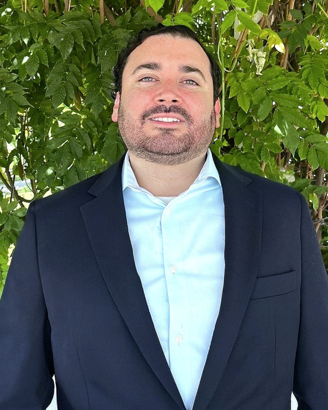 Man in a navy suit and light blue shirt smiling in front of green foliage.