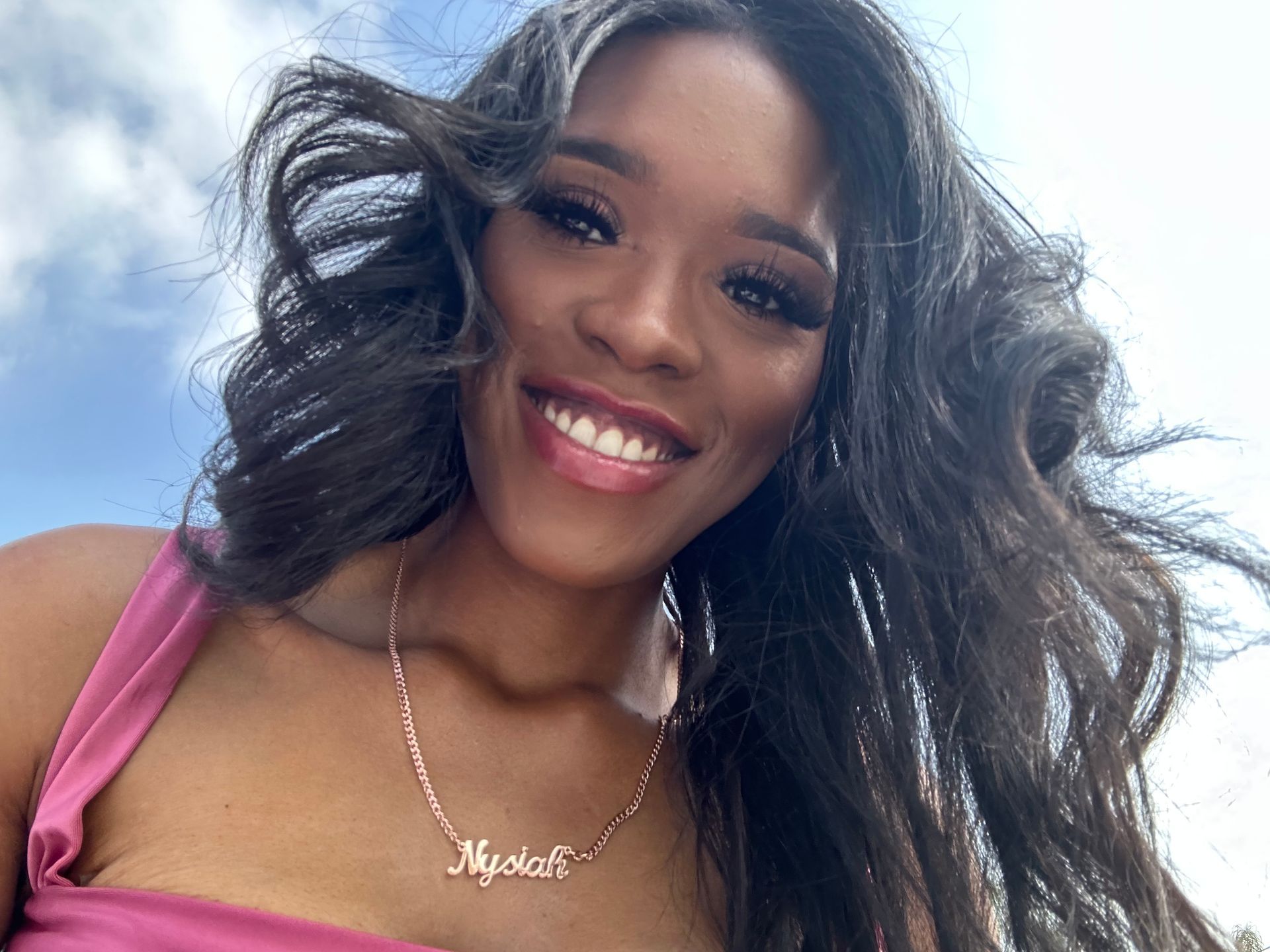 Woman smiles, wearing a pink top, rose gold necklace "Mystah". Dark wavy hair, outdoors, against a blue sky.