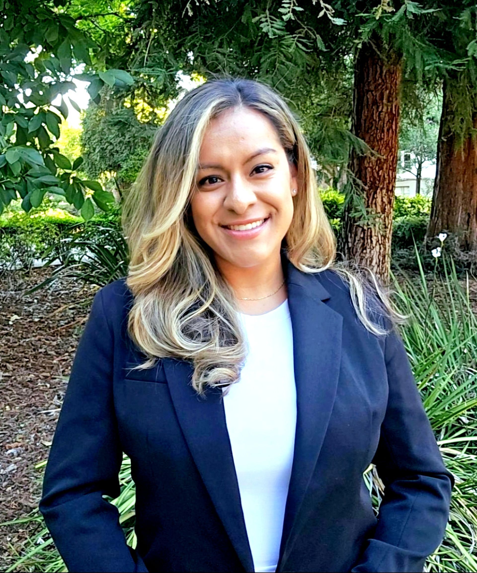A woman in a black jacket and white shirt is standing in front of trees.