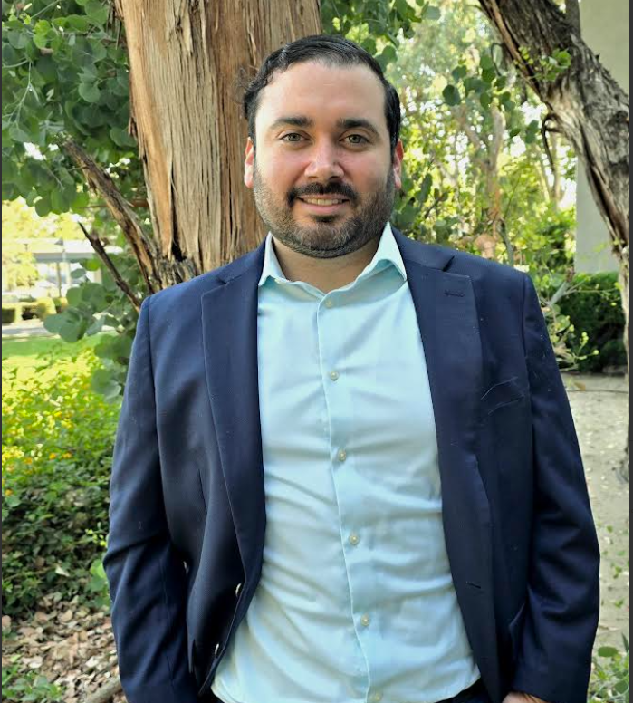 Man with a beard in a navy blazer and light blue shirt smiling, posing outdoors near a tree and greenery.
