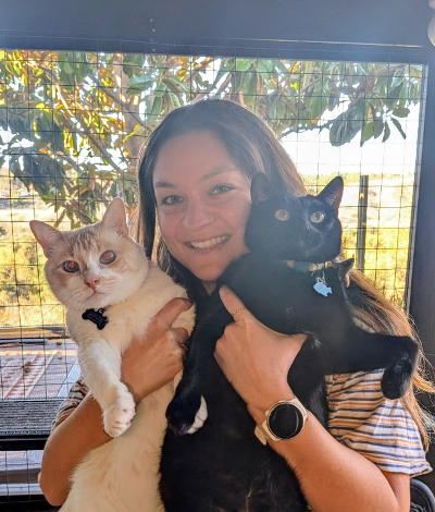 Woman smiles, holding a tan and a black cat outside, under a tree.