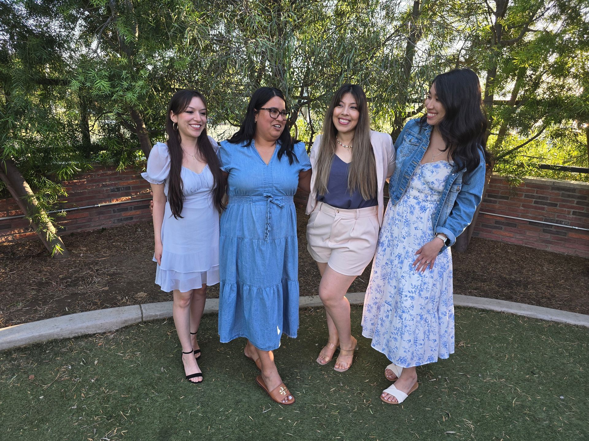 Four smiling people stand outdoors. Two wear blue dresses, one a denim jacket, and one a blazer over shorts and a top.