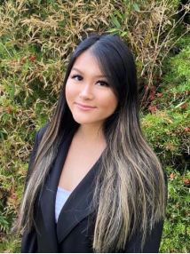 A woman in a black jacket is standing in front of a bush.