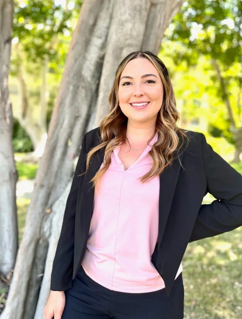 A woman in a pink shirt and black jacket is standing in front of a tree.