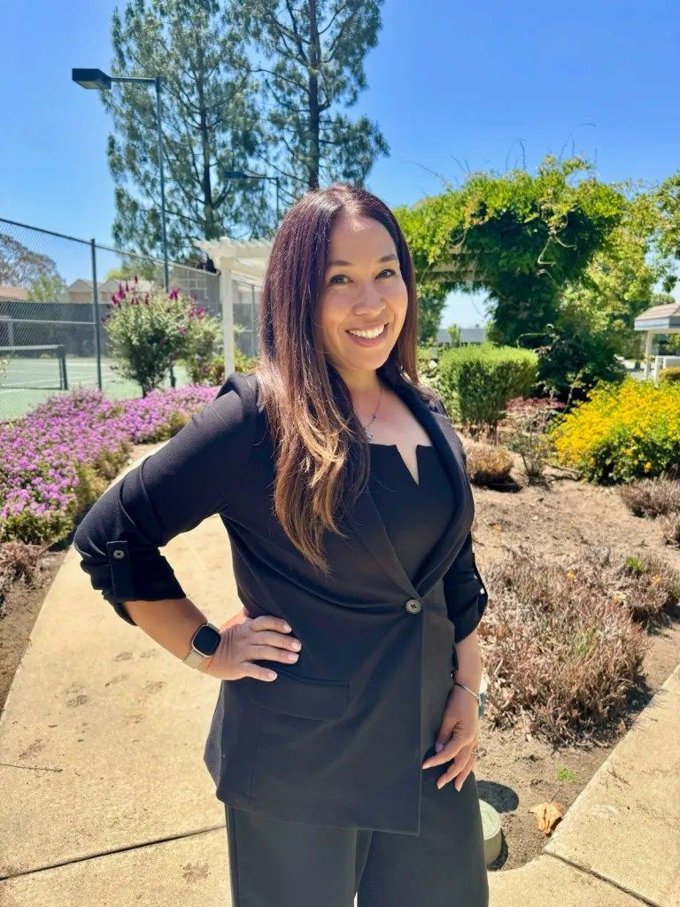 A woman in a black jacket is standing on a sidewalk with her hands on her hips.