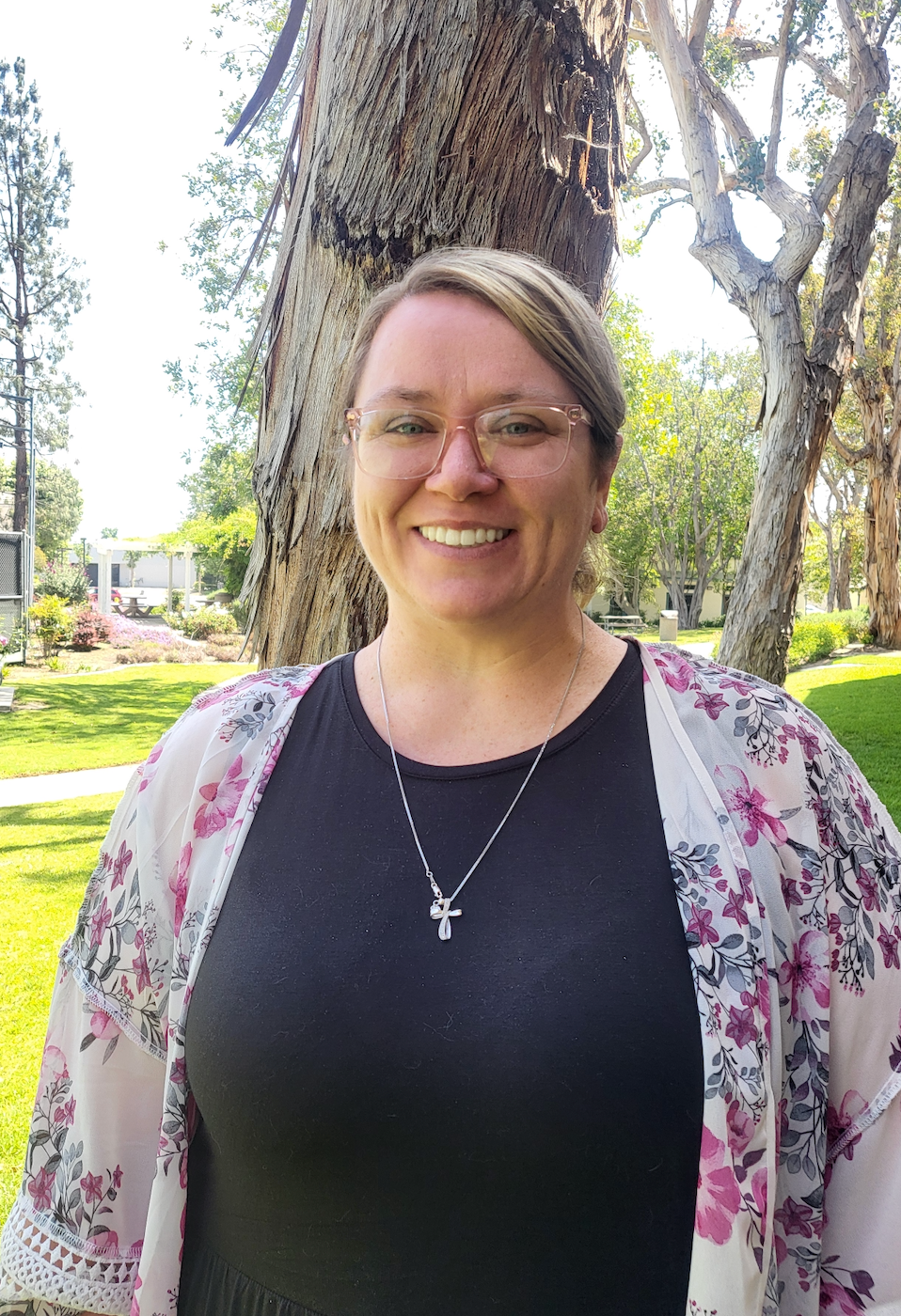 A woman wearing glasses and a floral kimono is smiling in front of a tree.