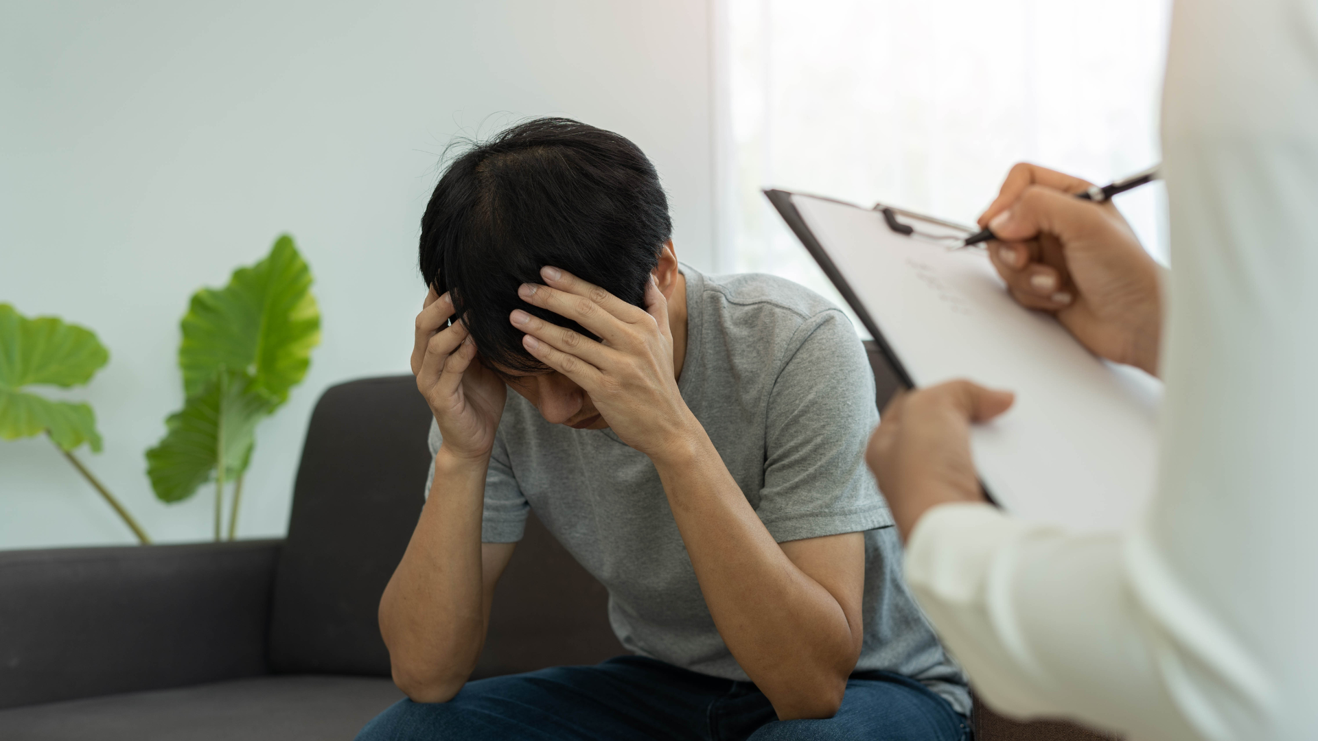 A man is sitting on a couch with his head in his hands while a doctor writes on a clipboard.