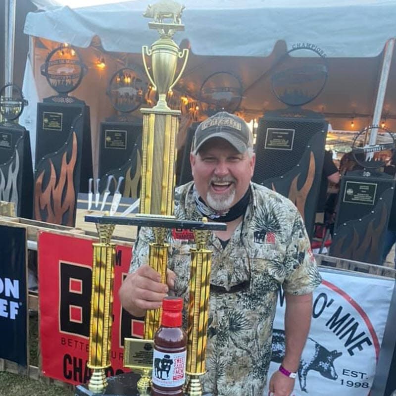 B&B Ambassador and Pitmaster Mark Lambert is holding a trophy on stage at a BBQ competition
