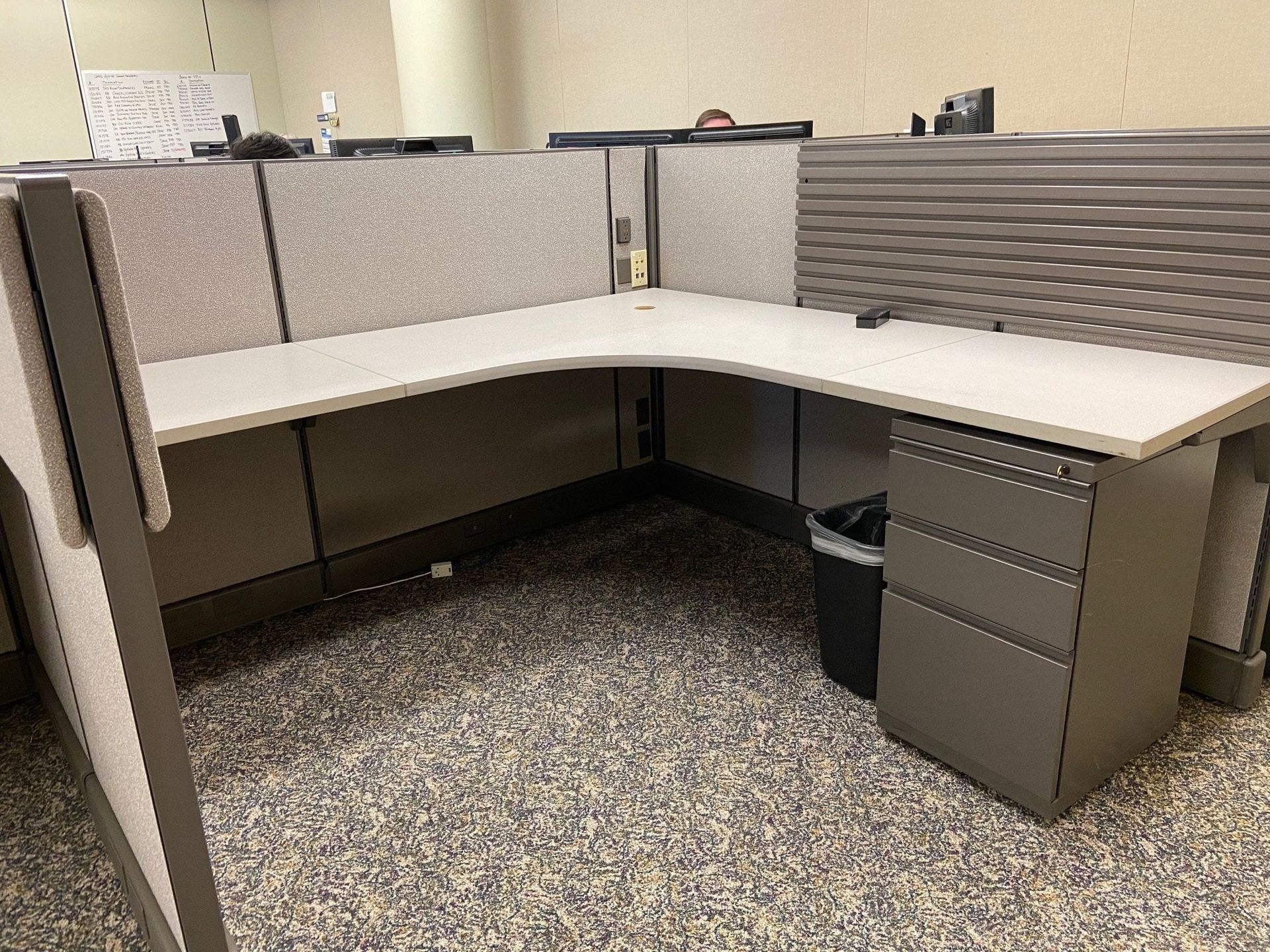 A cubicle with a desk , drawers , and a trash can.